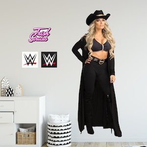 Può includere: Una donna indossa un cappello da cowboy nero, un cardigan nero a maniche lunghe, leggings neri e stivali neri. È in piedi davanti a un muro bianco con un logo rosa "Trish Stratus" e due loghi WWE in bianco e nero.