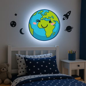May include: A cheerful, glowing Earth-shaped wall light with a smiling face, alongside black space-themed decals. The light is above a white bed with a navy blue star-patterned bedding set. A teddy bear is on the bed.