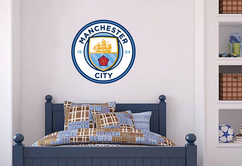 May include: A blue and white Manchester City Football Club logo decal with the text "Manchester City 1894" on a white wall above a blue bed with brown and blue patterned bedding.