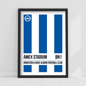 May include: Framed print featuring blue and white vertical stripes. The top left corner displays a circular logo. Below the stripes, the text reads "AMEX STADIUM BN1" and "BRIGHTON & HOVE ALBION FOOTBALL CLUB".