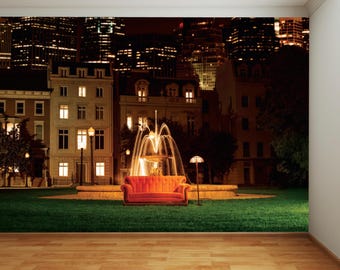 May include: A red orange sofa sits in front of a fountain in a city park at night. The city skyline is visible in the background.