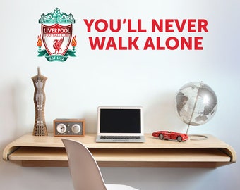 May include: A white wall with a red and green Liverpool Football Club crest and the text "You'll Never Walk Alone". The crest features a red bird with a white beak and a white football. The text is in a bold, red font. The image also shows a wooden desk with a laptop, a globe, and a red toy car.