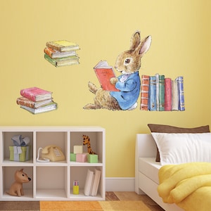Puede incluir: Una calcomanía de pared con un conejo con una chaqueta azul leyendo un libro, con pilas de libros a cada lado. El conejo está sentado sobre un fondo amarillo.