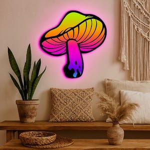 May include: A rainbow-coloured mushroom shaped wall light with a black outline. The light is mounted on a wall with a light brown wooden shelf below it. There is a potted plant, a throw pillow, and a vase with dried flowers on the shelf.