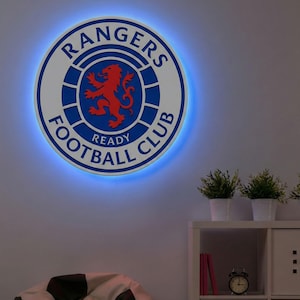 Op de afbeelding: Een rond, verlicht bord met het logo van de Rangers Football Club. Het bord is wit met blauwe en rode accenten, en de tekst "Rangers Football Club" is zichtbaar. Een blauw licht schijnt rond de rand van het bord.