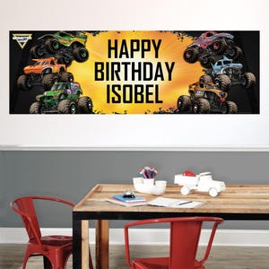 May include: A banner with the text "HAPPY BIRTHDAY ISOBEL" in large, bold, black letters. The banner features illustrations of monster trucks in various colors, including green, orange, red, and blue, against a yellow and black background.
