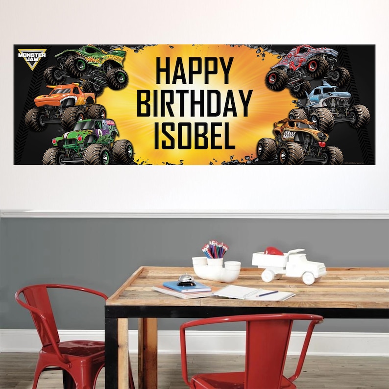 May include: A banner with the text "HAPPY BIRTHDAY ISOBEL" in large, bold, black letters. The banner features illustrations of monster trucks in various colors, including green, orange, red, and blue, against a yellow and black background.