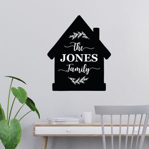 Op de afbeelding: Een zwarte, huisvormige wanddecoratie met de tekst "The Jones Family" in witte letters. Het ontwerp bevat witte bloemaccenten aan de boven- en onderkant. De decoratie is bevestigd aan een lichtgekleurde muur.