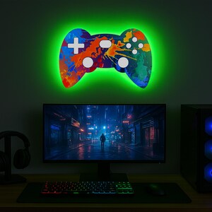 May include: A colourful, abstractly painted, illuminated game controller wall decor with a green glow. The controller is mounted on a wall above a computer monitor, keyboard and mouse.