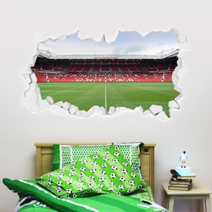 May include: A white wall with a hole in it revealing a green football pitch and a red stadium with the words "Manchester United" on the seats. A green and white football patterned bedspread and pillow are in the foreground.
