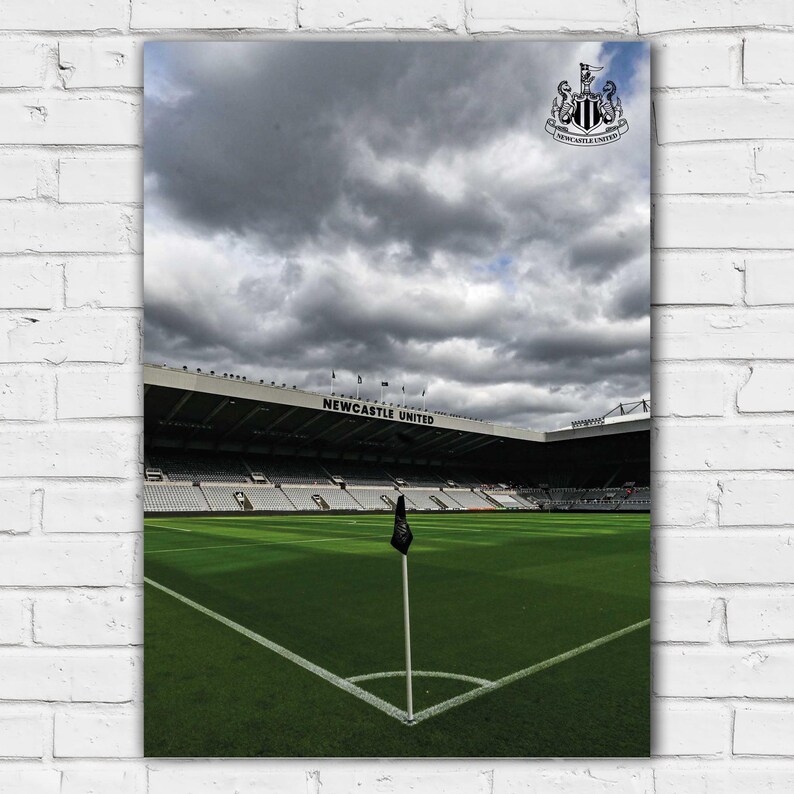May include: A black and white image of a soccer field with a corner flag. The field is surrounded by a large stadium with the words "NEWCASTLE UNITED" on the side. The sky is cloudy and gray.
