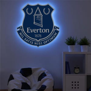 May include: A wall-mounted illuminated Everton Football Club crest in blue and white. The crest is shield-shaped, with the word "Everton" and the year "1878". The crest has a blue light outlining the edges, creating a glowing effect.