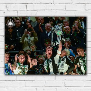 May include: A group of people celebrating with confetti and a silver trophy. The trophy has a green and white ribbon that says "Carabao Energy Cup Final". The people are wearing black and white uniforms and are holding the trophy up in the air.