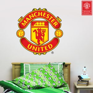 Op de afbeelding: Een rode en gele Manchester United voetbalteam logo sticker op een witte muur boven een bed met groene beddengoed en een voetbalpatroon.