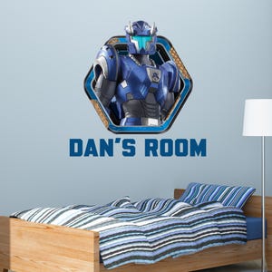 May include: A wall decal featuring a blue robot character within a hexagonal frame, with the text "DAN'S ROOM" below. The image is set against a light blue wall, with a wooden bed and striped bedding visible.