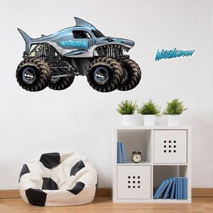 Op de afbeelding: Een grijze en blauwe monstertruck met een haaienontwerp, met het woord "MEGALODON" in blauw. De truck heeft oversized zwarte banden en staat tegen een witte achtergrond. Een bijpassende "MEGALODON" sticker is ook zichtbaar.