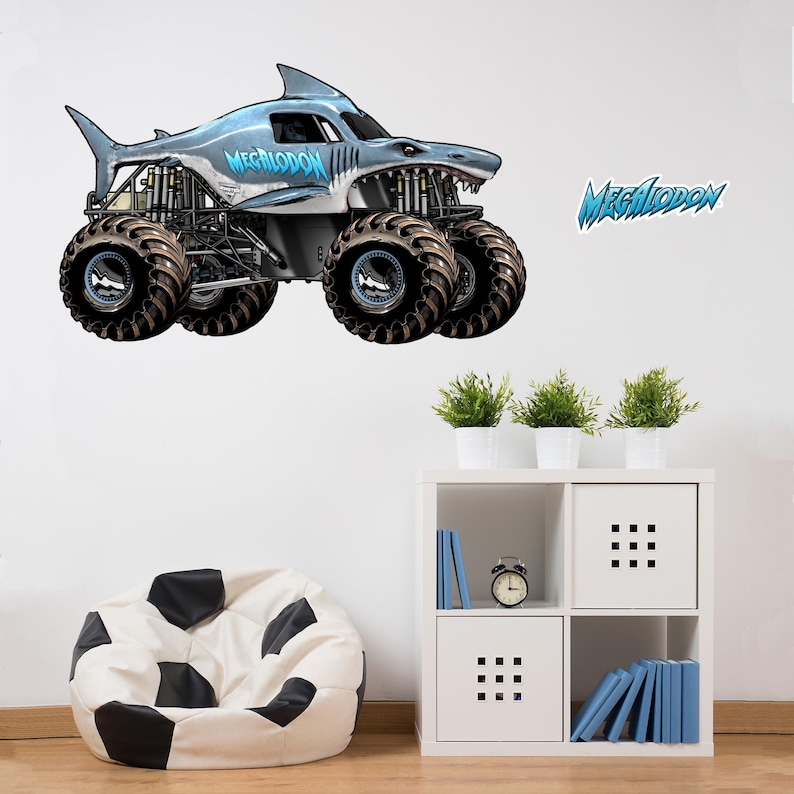 Op de afbeelding: Een grijze en blauwe monstertruck met een haaienontwerp, met het woord "MEGALODON" in blauw. De truck heeft oversized zwarte banden en staat tegen een witte achtergrond. Een bijpassende "MEGALODON" sticker is ook zichtbaar.