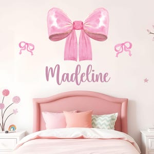 Op de afbeelding: Een muursticker set met een grote roze strik in aquarel-stijl, gecentreerd boven de naam Madeline in een paars cursief lettertype, geflankeerd door twee kleinere roze strikken, met extra kleine bloem- en stervormige accenten.
