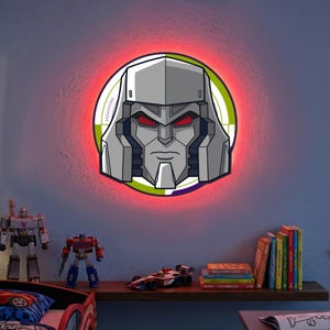 Pode incluir: Uma luminária de parede com o rosto de um personagem robô, com um esquema de cores cinza e preto e olhos vermelhos. A luz é cercada por um halo vermelho. A imagem é de um quarto infantil com brinquedos em uma prateleira.