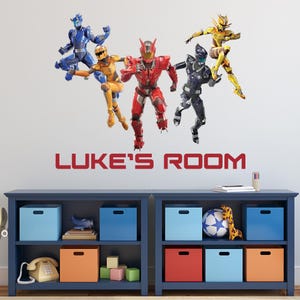 May include: A wall decal featuring five action figures in dynamic poses, each in a different color: blue, yellow, red, black, and gold. The text "LUKE'S ROOM" is displayed in red. Below, a blue bookshelf with storage boxes and decorative items.