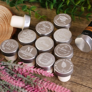May include: A set of 12 silver bottle stoppers with various engraved designs. The designs include names, dates, and phrases like "Eat Drink and Be Merry", "Cheers", "Love", and "Our Forever Begins Now".