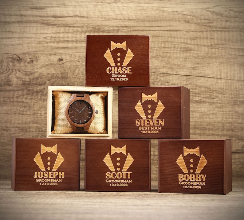 Op de afbeelding: Vijf houten dozen met gegraveerde smokingontwerpen en namen. De dozen zijn in een piramidevorm gerangschikt met een houten horloge in de middelste doos. De dozen zijn gelabeld met de namen Chase, Steven, Joseph, Scott en Bobby. De data 12.10.2026 zijn op elke doos gegraveerd.