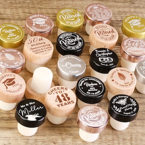 May include: Assortment of personalized bottle stoppers with various designs and text, including graduation, wedding anniversary, birthday, and retirement. The stoppers are made of wood, metal, and cork.