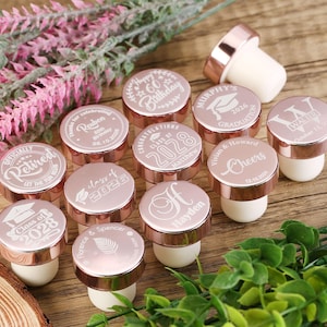 May include: Rose gold bottle stoppers with various engraved designs, including graduation caps, names, and the year 2028. The stoppers are arranged on a wooden surface.