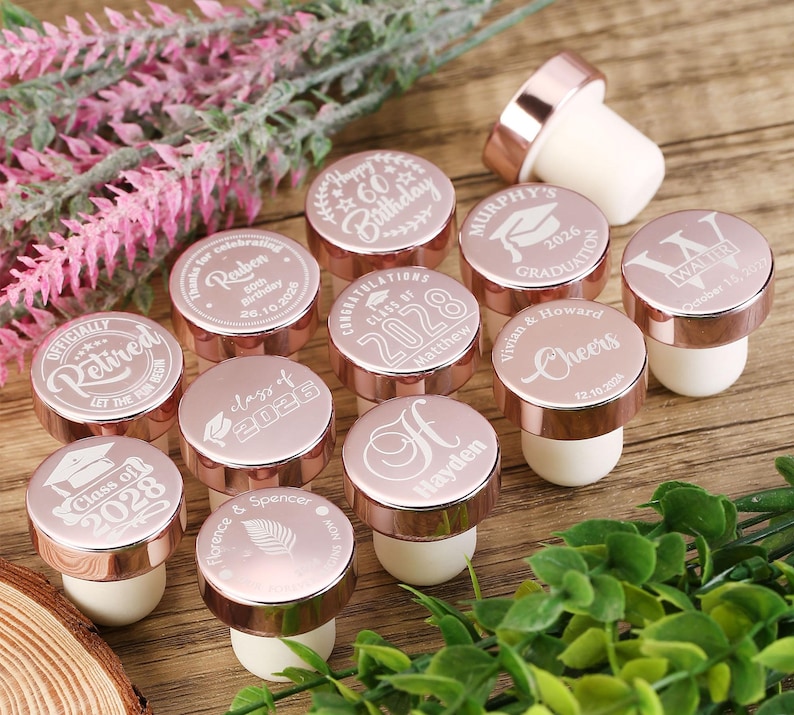 May include: Rose gold bottle stoppers with various engraved designs, including graduation caps, names, and the year 2028. The stoppers are arranged on a wooden surface.
