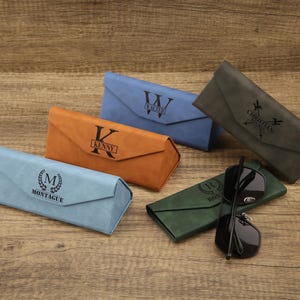 May include: An assortment of personalized eyeglass cases in various colors, including blue, orange, and green. Each case features a unique name or initial, such as "Walter" and "Kenny", and a pair of black sunglasses.