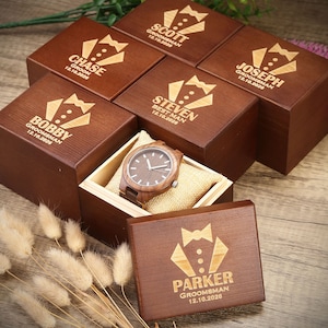 Op de afbeelding: Een set houten dozen met gegraveerde tekst, elke doos bevat een houten polshorloge. De dozen zijn bruin en hebben een strikdasontwerp. De tekst op de dozen bevat namen en data. De dozen zijn in een cirkel op een houten oppervlak gerangschikt.