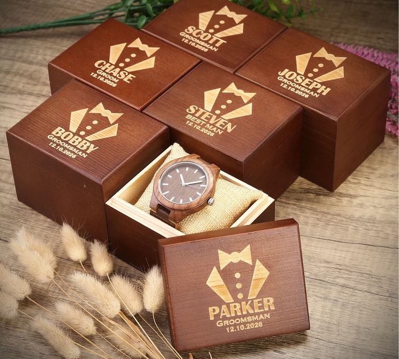 Op de afbeelding: Een set houten dozen met gegraveerde tekst, elke doos bevat een houten polshorloge. De dozen zijn bruin en hebben een strikdasontwerp. De tekst op de dozen bevat namen en data. De dozen zijn in een cirkel op een houten oppervlak gerangschikt.