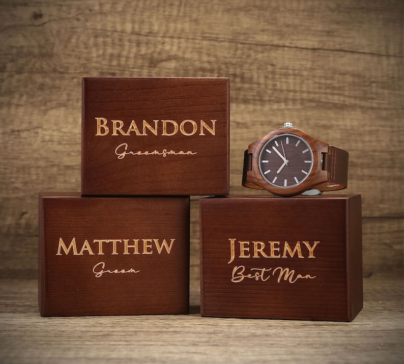 Op de afbeelding: Drie houten dozen met gegraveerde namen en titels: Brandon -  Groomsman, Matthew - Groom, en Jeremy - Best Man. Een houten polshorloge met een bruine wijzerplaat en een bruin leren bandje ligt op de dozen.