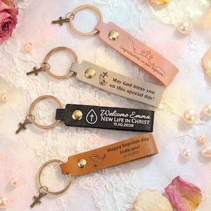 May include: Four leather keyrings with gold-coloured hardware and cross charms. The keyrings are in pink, beige, black, and tan. Each keyring has a different message, including "Congratulations on your baptism!" and "Welcome Emma NEW LIFE IN CHRIST 11.10.2029."