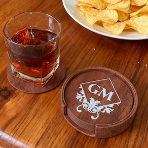 Puede incluir: Un juego de posavasos de madera marrón con un monograma blanco y un diseño decorativo. Un vaso de líquido ámbar se encuentra sobre un posavasos a juego. Un plato de patatas fritas está en el fondo.