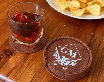 May include: A set of brown wooden coasters with a white monogram and decorative design. A glass of amber liquid sits on a matching coaster. A plate of potato chips is in the background.