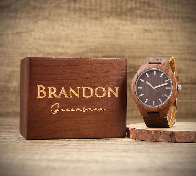 Op de afbeelding: Een houten horloge met een bruin leren bandje in een houten doos. De doos is gegraveerd met de naam "BRANDON" en het woord "Groomsman".