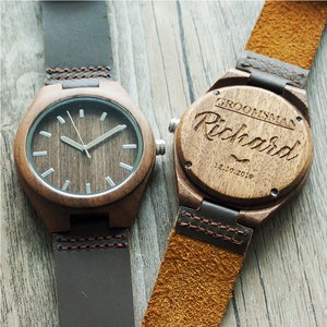 Op de afbeelding: Twee houten polshorloges met bruine leren bandjes. Een horloge heeft een witte wijzerplaat met zwarte wijzers. De andere horloge heeft de woorden "Groomsman Richard 12.10.2018" gegraveerd.