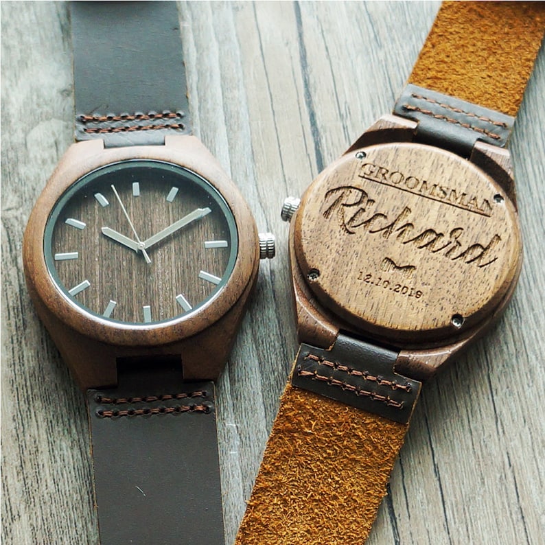 Op de afbeelding: Twee houten polshorloges met bruine leren bandjes. Een horloge heeft een witte wijzerplaat met zwarte wijzers. De andere horloge heeft de woorden "Groomsman Richard 12.10.2018" gegraveerd.