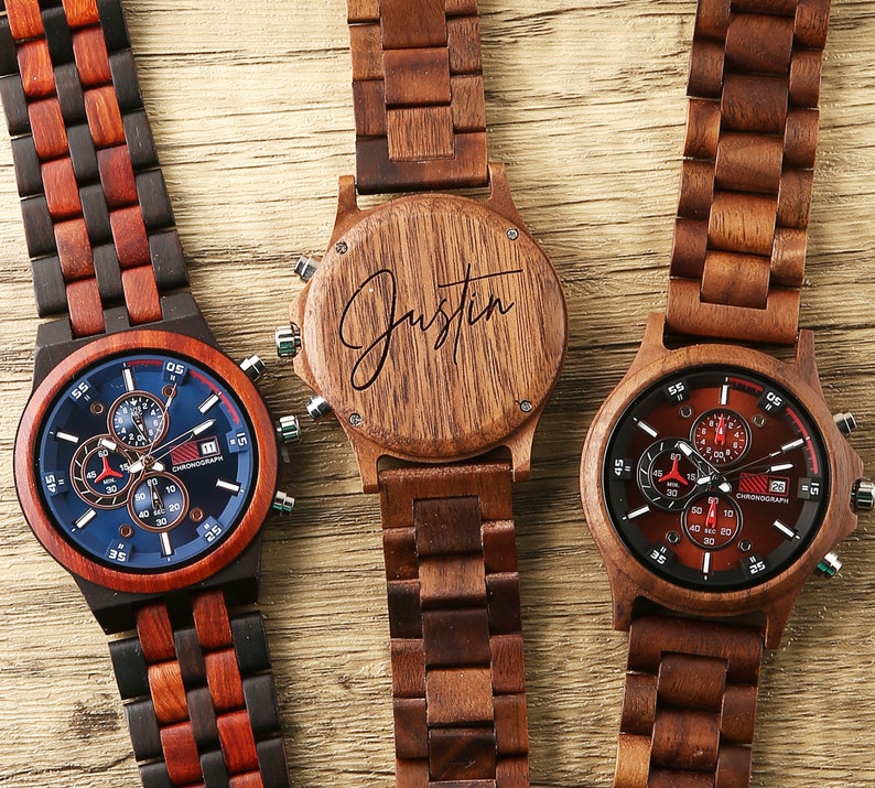 May include: Three wooden wristwatches with chronograph faces. The watch on the left has a blue face with red accents. The watch in the middle has a wooden face with the name "Justin" engraved on it. The watch on the right has a red face with black accents.