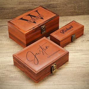 Può includere: Tre scatole di legno con coperchi in pelle. Le scatole sono incise con nomi e date. La scatola più grande ha inciso "Walter". La scatola più piccola ha inciso "Richard 12.10.2028". La scatola media ha inciso "Justin GROOMSMAN".