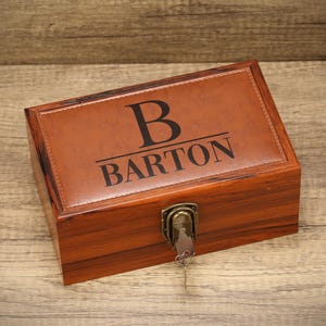 May include: A rectangular wooden box with a brown leather top. The top is embossed with the letter "B" above the name "BARTON" in black. The box has a metal clasp and a key.