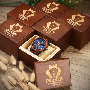 May include: A wooden box with a bow tie design and the text "Parker Groomman 12.10.2026" engraved on the lid. The box is surrounded by other similar boxes with different names and dates engraved on them. A wooden watch with a blue face and brown leather band is in a separate box.