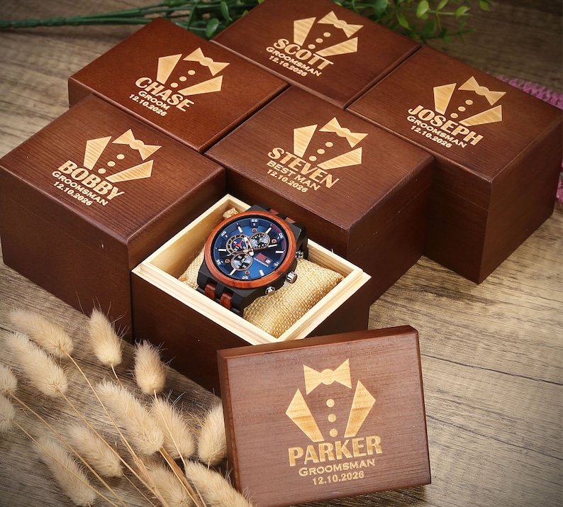 May include: A wooden box with a bow tie design and the text "Parker Groomman 12.10.2026" engraved on the lid. The box is surrounded by other similar boxes with different names and dates engraved on them. A wooden watch with a blue face and brown leather band is in a separate box.