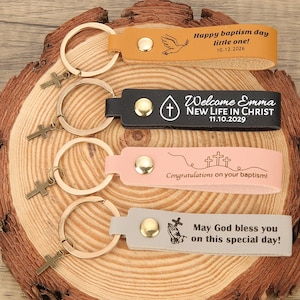 May include: Four leather keyrings with gold-tone rings and cross charms. The keyrings are in tan, black, pink, and grey. Each keyring has a different message: "Happy baptism day little one!", "Welcome Emma NEW LIFE IN CHRIST", "Congratulations on your baptism!", and "May God bless you on this special day!"