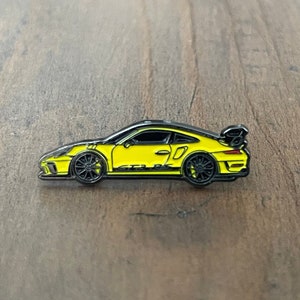 May include: A yellow sports car pin with black accents and wheels. The pin features the text "GT3 RS" on the side and a black spoiler. The car is set against a wooden background.