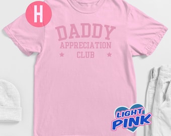 May include: Light pink t-shirt with the text "DADDY APPRECIATION CLUB" in a matching pink color. The design includes two stars. The shirt is a crew neck style.