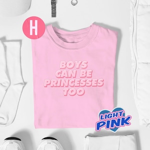 Puede incluir: Una camiseta rosa claro con el texto "BOYS CAN BE PRINCESSES TOO" en una fuente rosa en capas. La camiseta está rodeada de accesorios blancos, incluyendo un gorro, calcetines y zapatillas. Una pegatina dice "LIGHT PINK".