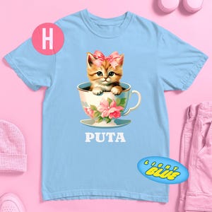 May include: Light blue t-shirt featuring a cartoon kitten with a pink bow sitting in a teacup with floral designs. The word "PUTA" is printed in white below the teacup. A pink circle with the letter "H" is on the left.