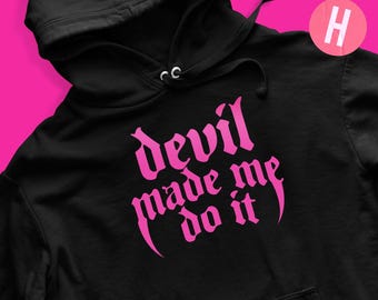 Könnte beinhalten: Schwarzer Kapuzenpullover mit Fronttasche und Kordelzugkapuze. Der Pullover zeigt den pinkfarbenen Text "devil made me do it" in einer gotischen Schrift. Das Bild enthält auch einen pinkfarbenen und blauen Aufkleber mit der Aufschrift "OPTION 1" und einen pinkfarbenen Kreis mit dem Buchstaben "H".